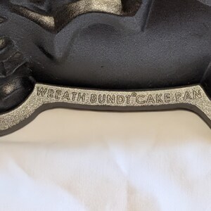 May include: Close-up of a black Wreath Bundt Cake Pan with a silver rim. The pan has a decorative design and is made of cast iron. The text "WREATH BUNDT CAKE PAN" is embossed on the rim.