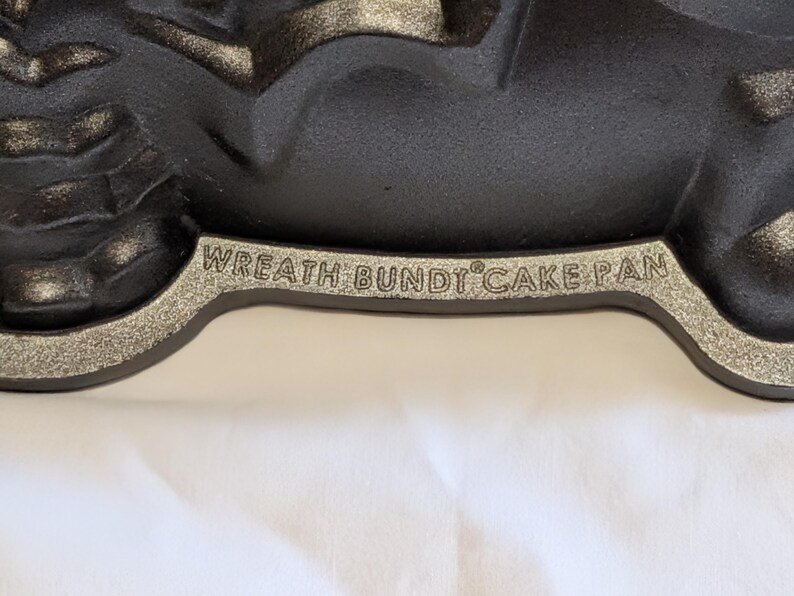 May include: Close-up of a black Wreath Bundt Cake Pan with a silver rim. The pan has a decorative design and is made of cast iron. The text "WREATH BUNDT CAKE PAN" is embossed on the rim.