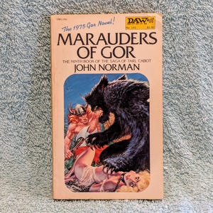 May include: A paperback book cover with a pink background and the title "Marauders of Gor" in black text. The book is the ninth in a series and is written by John Norman. The cover art depicts a woman in a white dress being attacked by a large black bear. A man with a sword is trying to help her.