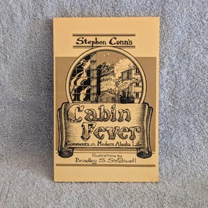 May include: A tan book cover with a black and white illustration of a city skyline. The title of the book is "Cabin Fever: Comments on Modern Alaska Life" by Stephen Conn's. The book cover also includes the text "Illustrations by Bradley S. Stockwell".