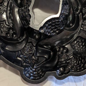 May include: A dark gray, decorative bundt cake pan with a wreath design. The pan features detailed embossed patterns of berries, leaves, and spiderwebs. The center of the pan is a smooth, unadorned circle.