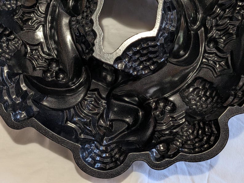 May include: A dark gray, decorative bundt cake pan with a wreath design. The pan features detailed embossed patterns of berries, leaves, and spiderwebs. The center of the pan is a smooth, unadorned circle.