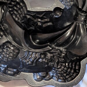 May include: A dark gray, intricately designed bundt pan with a decorative pattern. The pan features detailed relief designs of pinecones, leaves, and other festive elements. The pan's surface has a metallic sheen, suggesting a non-stick coating.