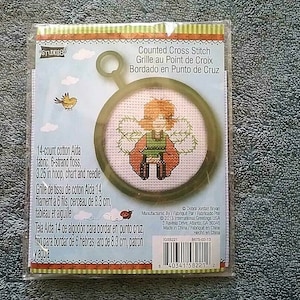 May include: A counted cross stitch kit featuring a green and red fairy with a yellow bird on a white background. The kit includes 14-count Aida fabric, 6-strand floss, a 3.25 inch hoop, chart and needle. The kit is manufactured by Debra Jordan Bryan.