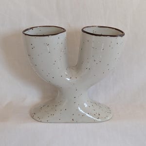 May include: A speckled, off-white ceramic vase with two openings. The vase has a unique Y-shape, with a rounded base and dark brown trim around the openings. The surface is dotted with small brown specks.