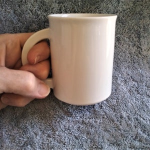 K&ouml;nnte beinhalten: Eine wei&szlig;e Keramiktasse mit Henkel. Die Tasse steht auf einer grauen, strukturierten Oberfl&auml;che.
