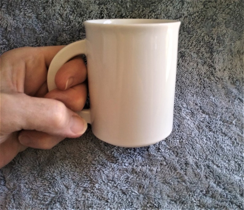 K&ouml;nnte beinhalten: Eine wei&szlig;e Keramiktasse mit Henkel. Die Tasse steht auf einer grauen, strukturierten Oberfl&auml;che.