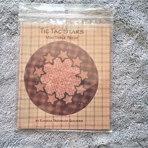 May include: A brown and white printed pattern with a star design. The pattern is titled "Tic Tac Stars Mini Table Treat" and is by "Kansas Troubles Quilters".
