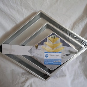 May include: A set of three square, silver-colored baking pans, nested together. The pans are packaged in clear plastic wrap. A cardboard insert displays a cake and the text "3-Tier Diamond Pan Set".