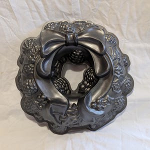 May include: A dark gray, decorative Bundt cake pan in the shape of a wreath. The pan features a large bow at the top and detailed floral and berry designs around the edges. The pan is made of metal.