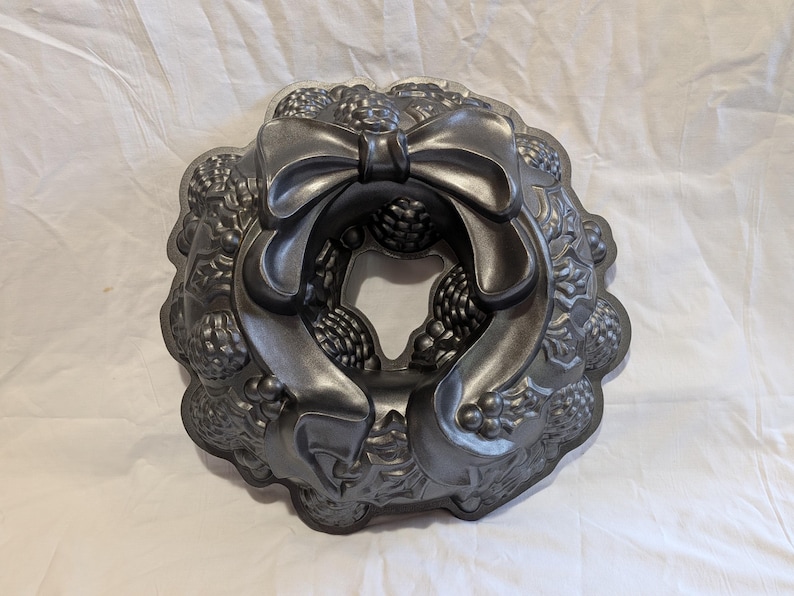 May include: A dark gray, decorative Bundt cake pan in the shape of a wreath. The pan features a large bow at the top and detailed floral and berry designs around the edges. The pan is made of metal.