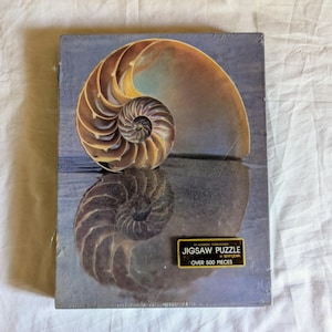 May include: A jigsaw puzzle box featuring a nautilus shell design. The shell is depicted in shades of brown, yellow, and tan, with a mirrored reflection below. The box has a rectangular shape and includes text that reads "Jigsaw Puzzle Over 500 Pieces."