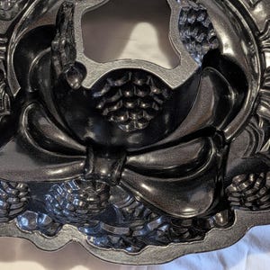 May include: A dark gray, decorative bundt cake pan with a detailed design. The pan features a bow, pinecones, and floral accents. The pan has a scalloped edge and is made of a metallic material.