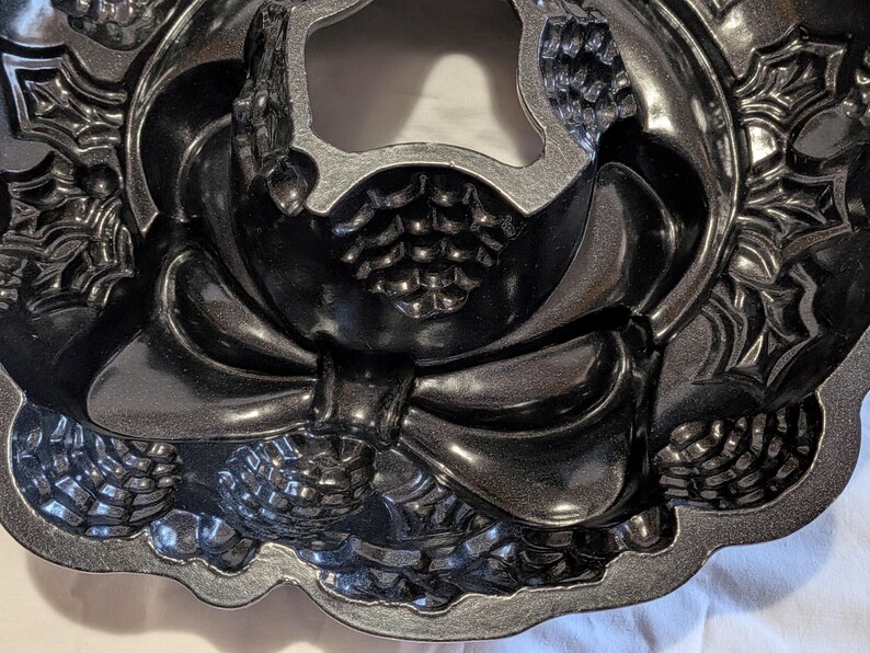 May include: A dark gray, decorative bundt cake pan with a detailed design. The pan features a bow, pinecones, and floral accents. The pan has a scalloped edge and is made of a metallic material.