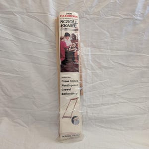 May include: A rectangular package for a scroll frame, with the text "SCROLL FRAME" in large letters. The package features an illustration of people working on a craft project. The package is labeled "2999 F.A. EDMUNDS" and lists "Cross Stitch, Needlepoint, Crewel, Embroidery."