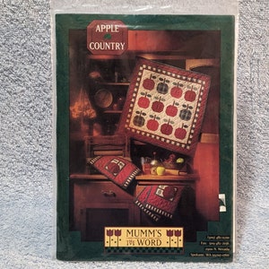 May include: A quilt pattern featuring a red apple design with green leaves. The pattern is called "Apple Country" and is from "Mumm's the Word".