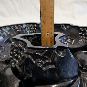May include: A black, ornate bundt pan with a decorative floral and grape design. A wooden ruler is placed inside the pan, showing a depth of approximately 4 inches. The pan is made of a shiny, dark material.