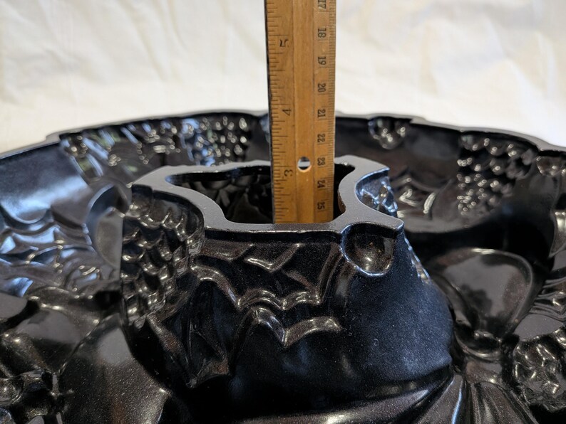 May include: A black, ornate bundt pan with a decorative floral and grape design. A wooden ruler is placed inside the pan, showing a depth of approximately 4 inches. The pan is made of a shiny, dark material.
