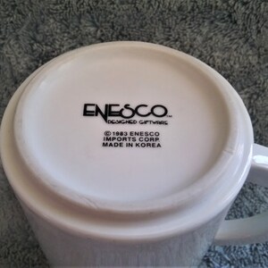 K&ouml;nnte beinhalten: Wei&szlig;e Keramiktasse mit einem schwarzen Logo, auf dem "Enesco Designed Giftware" und "&copy; 1983 Enesco Imports Corp. Made in Korea" steht.