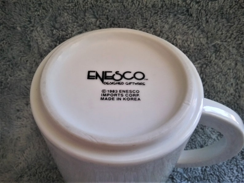 K&ouml;nnte beinhalten: Wei&szlig;e Keramiktasse mit einem schwarzen Logo, auf dem "Enesco Designed Giftware" und "&copy; 1983 Enesco Imports Corp. Made in Korea" steht.