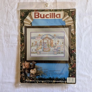 May include: A Bucilla counted cross-stitch kit featuring a framed nativity scene. The artwork depicts the birth of Jesus with angels, animals, and figures in soft pastel colors. The kit includes the text "Nativity" and "14" x 16" Counted Cross-Stitch Picture".