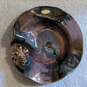 May include: A dark purple and blue ceramic bowl with a gold-colored design on the side. The bowl has a wavy edge and is handmade.