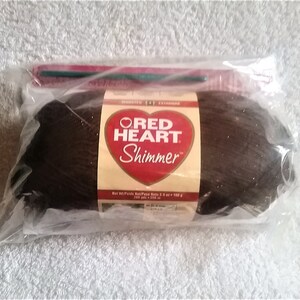 May include: A package of Red Heart Shimmer yarn in a dark brown color. The label features a red heart with the brand name and product details. A green crochet hook is visible inside the clear plastic packaging.