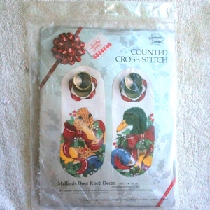 May include: A sealed package containing a counted cross-stitch kit. The kit features a design for door knob decor with two mallard duck patterns, each surrounded by festive bows and holly. The text "Mallards Door Knob Decor" is visible.
