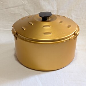 May include: A round, metallic gold cooking pot with a matching lid. The lid features a black, flower-shaped knob at the center and a series of small, rectangular ventilation slots arranged in a circular pattern around the top surface.