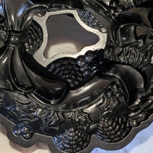 May include: A dark gray, intricately designed Bundt cake pan. The pan features a wreath-like pattern with pinecones, bows, and other decorative elements. The center of the pan has a unique, irregular shape. The pan is made of a metallic material.