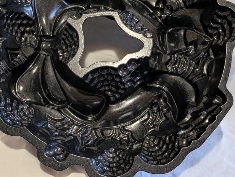 May include: A dark gray, intricately designed Bundt cake pan. The pan features a wreath-like pattern with pinecones, bows, and other decorative elements. The center of the pan has a unique, irregular shape. The pan is made of a metallic material.