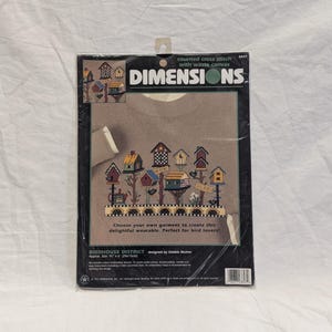 May include: A Dimensions counted cross stitch kit featuring a birdhouse design. The kit includes a brown sweatshirt with a colorful birdhouse pattern, the text "BIRDHOUSE DISTRICT", and the text "Choose your own garment to create this delightful wearable."