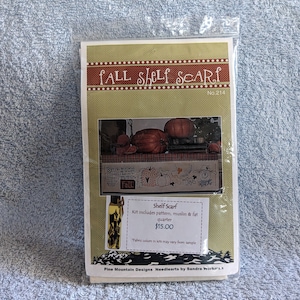 May include: A craft kit for a fall-themed shelf scarf. The kit includes a pattern, muslin fabric, and a fat quarter. The image shows a sample of the finished project, featuring pumpkins, gourds, and a scarecrow. The kit costs $15.00.