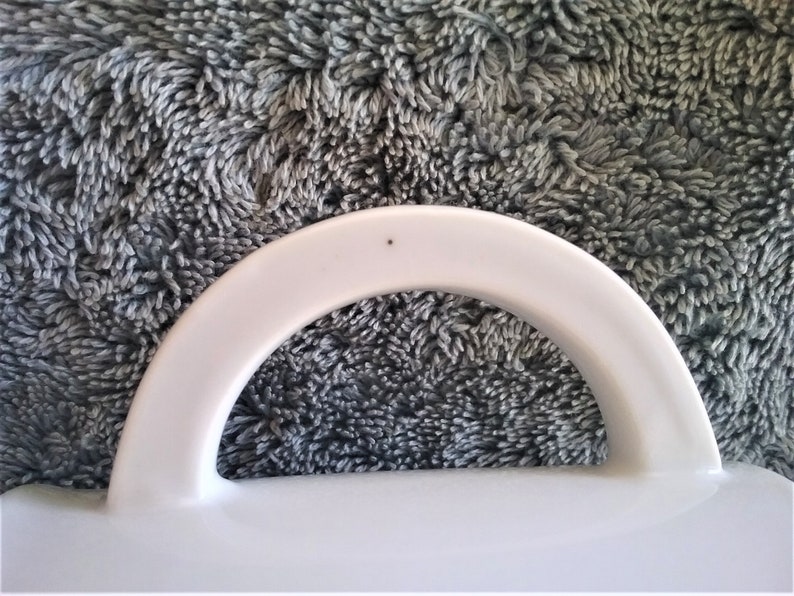 K&ouml;nnte beinhalten: Wei&szlig;er Keramikgriff mit einem kleinen schwarzen Punkt oben. Der Griff ist gebogen und hat eine glatte Oberfl&auml;che.