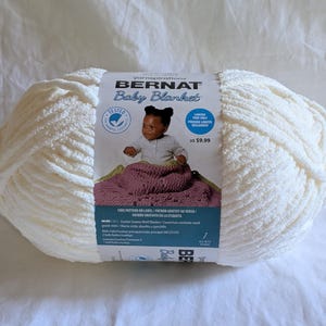 May include: A large skein of off-white Bernat Baby Blanket yarn. The label features a baby and a crocheted blanket. The yarn is a soft, plush material, ideal for baby blankets and other cozy projects.
