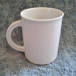 K&ouml;nnte beinhalten: Eine schlichte wei&szlig;e Keramiktasse mit Henkel. Die Tasse steht auf einer grauen, strukturierten Oberfl&auml;che.