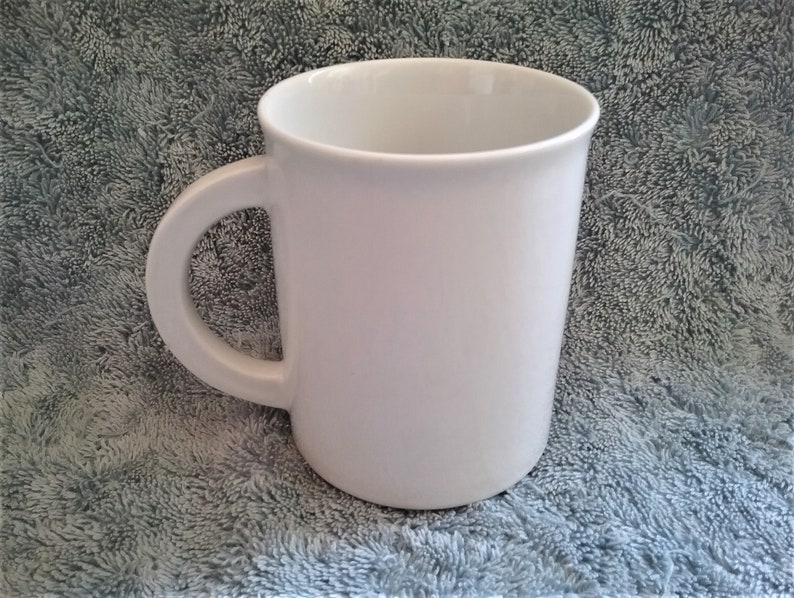 K&ouml;nnte beinhalten: Eine schlichte wei&szlig;e Keramiktasse mit Henkel. Die Tasse steht auf einer grauen, strukturierten Oberfl&auml;che.
