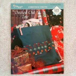 May include: A teal tote bag with a cross-stitch design of Christmas trees and houses. The bag is filled with wrapped gifts and candy canes. The text "Decked Out Tote" is printed on the bag. The text "Christmas Visions" is printed at the top of the image.