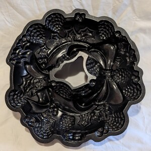 May include: Black cast iron bundt pan with a wreath design. The pan features detailed embossed patterns of pinecones, leaves, and bows. The center of the pan has a decorative shape. Ideal for baking festive cakes.