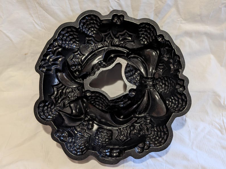 May include: Black cast iron bundt pan with a wreath design. The pan features detailed embossed patterns of pinecones, leaves, and bows. The center of the pan has a decorative shape. Ideal for baking festive cakes.