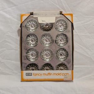 May include: A vintage Cooks Tools fancy muffin mold pan. The metal pan has 12 flower-shaped molds. The box is orange and white with the words "fancy muffin mold pan" printed on the front.