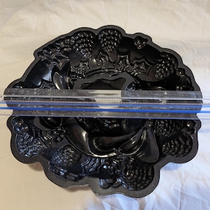 May include: A black, decorative cake pan with a detailed, embossed design featuring grapes, pinecones, and other festive elements. The pan is approximately 10 inches in diameter, perfect for baking holiday treats.