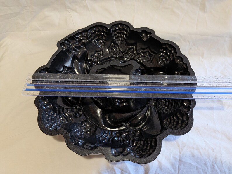 May include: A black, decorative cake pan with a detailed, embossed design featuring grapes, pinecones, and other festive elements. The pan is approximately 10 inches in diameter, perfect for baking holiday treats.