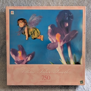 May include: A 750 piece jigsaw puzzle featuring a fairy with purple wings flying over a field of purple crocuses. The puzzle box has a pink background with the artist's name, Valerie Tabor-Smith, and the title "750 Piece Puzzle".