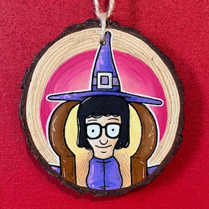 May include: A round wooden ornament featuring a cartoon character wearing a purple witch hat and glasses. The character is set against a pink and yellow background, with a brown chair. The ornament is suspended by a natural twine.