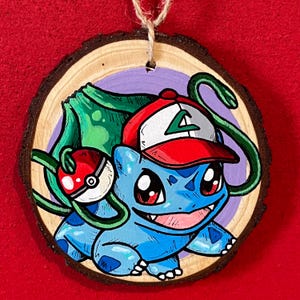 Könnte beinhalten: Handbemalter Holzschmuck mit einer blauen Cartoonfigur, die eine rot-weißes Kappe trägt. Die Figur hat rote Augen und einen lächelnden Ausdruck. Eine grüne Ranke und ein rot-weißer Ball sind ebenfalls abgebildet. Der Anhänger ist rund mit einem natürlichen Holzrand.