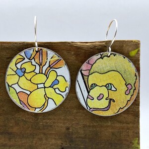 May include: A pair of round, silver-toned earrings. One earring features a yellow floral design with blue and orange accents. The other earring displays a cartoon-style yellow face with pink and green details. Both earrings have silver hooks.