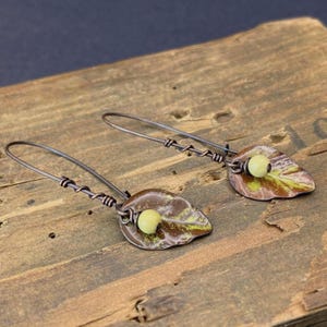 May include: A pair of dangle earrings with a leaf-shaped charm and a small yellow bead. The earrings are made of copper wire and have a rustic, vintage look.