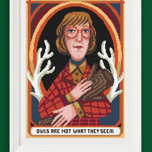 Può includere: Uno schema di punto croce con una donna con capelli biondi corti che indossa occhiali e una camicia a quadri rossi e neri. Tiene un tronco marrone tra le braccia. Lo sfondo è blu scuro con un disegno di corna di cervo bianche. Il testo "OWLS ARE NOT WHAT THEY SEEM" è in basso nell'immagine.