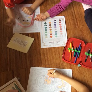 May include: Three children are coloring in a room with wooden floors. They are using colored pencils and a color guide. The color guide has 36 different colors. The children are coloring in a variety of images, including a girl with a crown, a girl with a flamingo, and a girl with a flower.